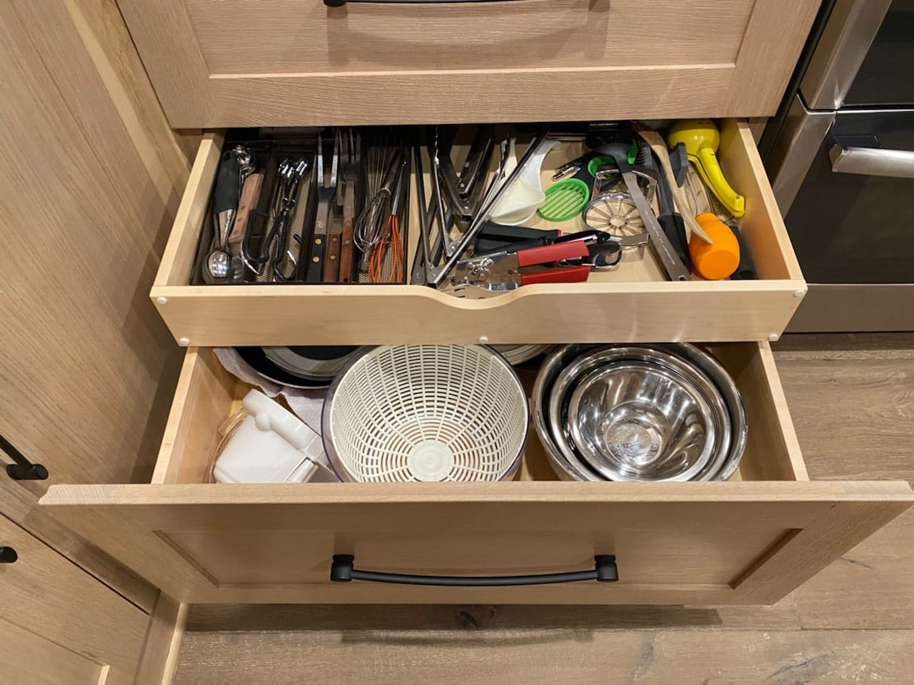Open deep drawers in custom white oak kitchen cabinets holding bowls and utensils
